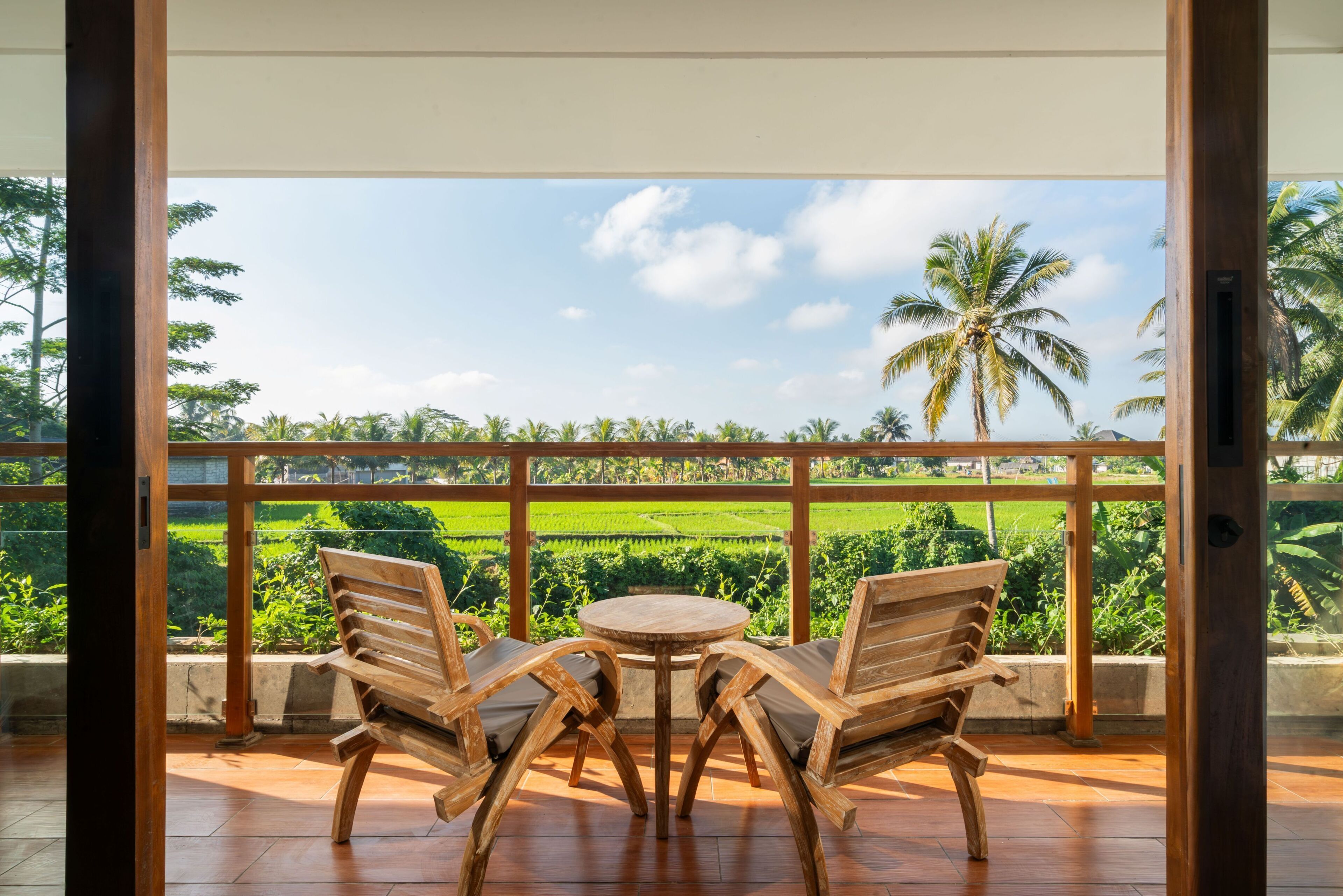 A wooden bench and table on a balcony with a view of green fields and palm trees.