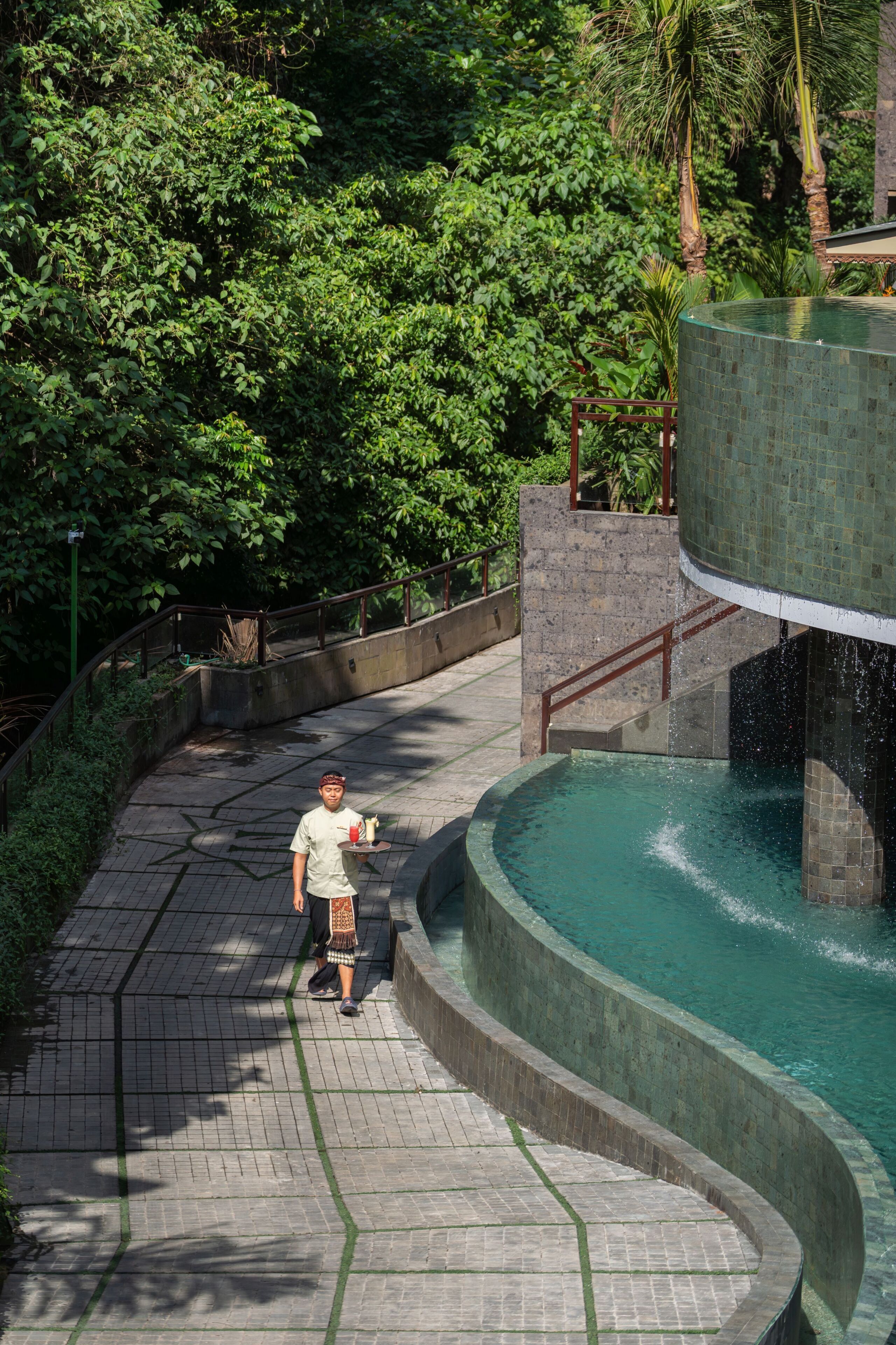 A man walking on a tiled pathway beside a pool with a water feature.