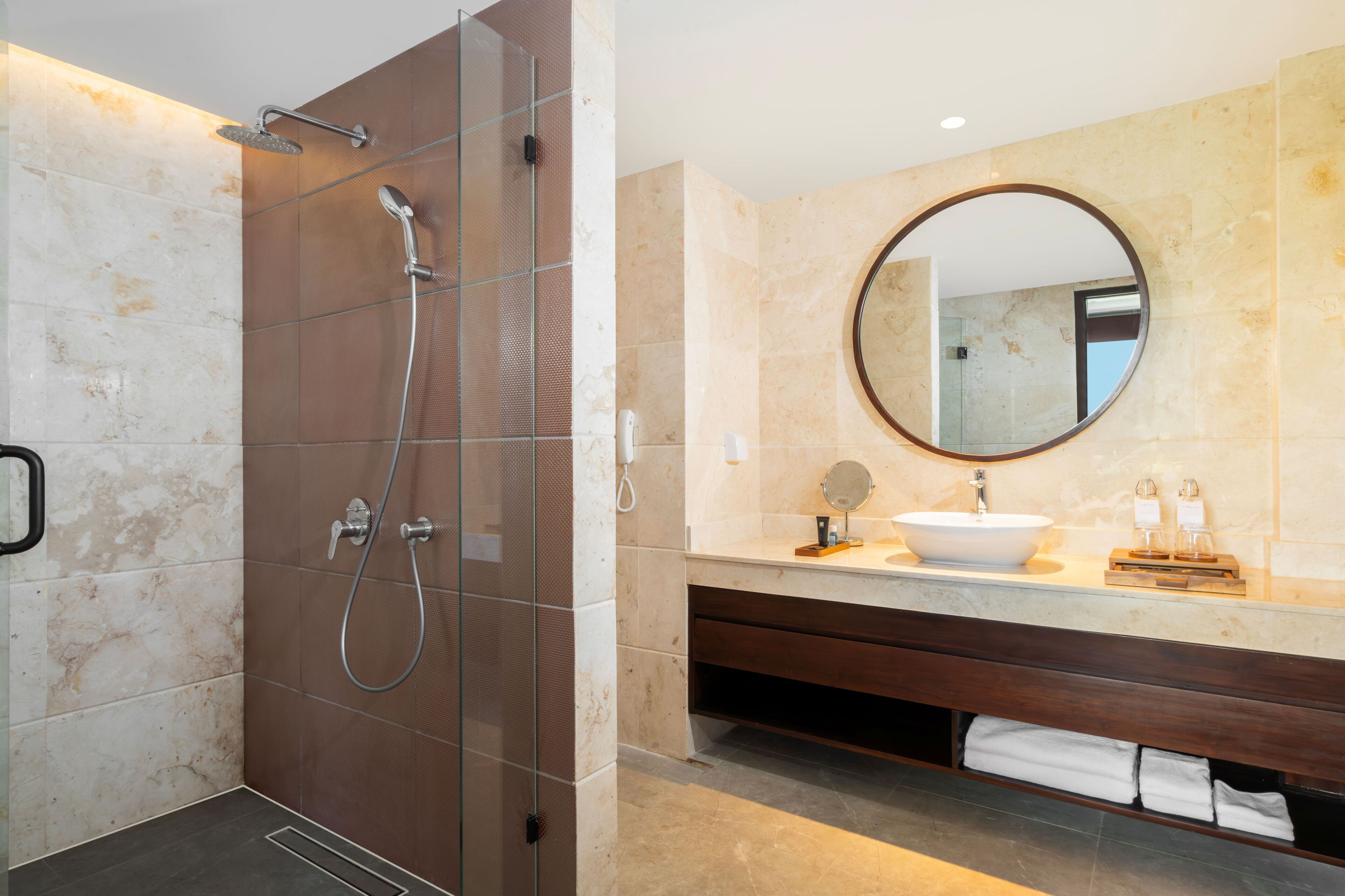 A modern bathroom with a glass-enclosed shower, a round mirror, a sink, and a wooden vanity.