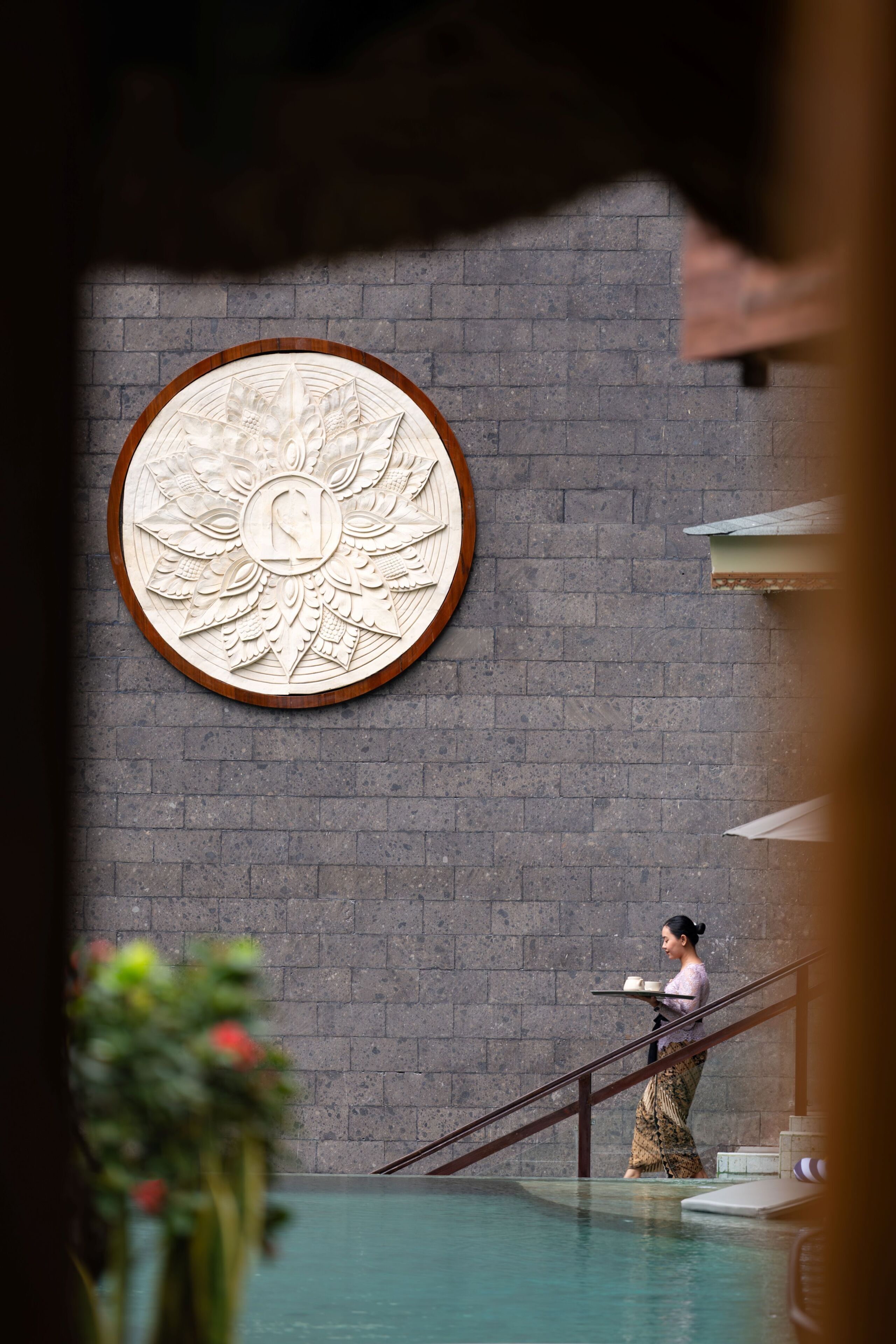 A person carrying trays near a pool with a large circular wall decoration.