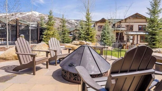 Zona de estar al aire libre con sillas Adirondack y una chimenea, rodeada de vistas a la montaña y un edificio con techo inclinado.
