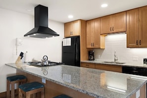 Eine moderne Küche mit Granit Arbeitsplatten, Holzschränken, einer schwarzen Dunstabzugshaube, einem schwarzen Kühlschrank und einer Kaffeemaschine.