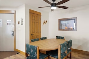 Ein Essbereich mit einem runden Holztisch, grünen Polsterstühlen, einem Deckenventilator, einer Holztür und einem gerahmten Bild an der Wand.