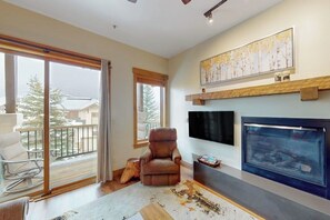 A living room with a fireplace, a large window, and a balcony. - Walk-to-lift condo w/ shared hot tubs/pool/gym & amazing mountain views (Steamboat Springs)