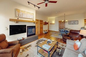 A living room with a fireplace, a television, a ceiling fan, and a dining area with a table and chairs. - Walk-to-lift condo w/ shared hot tubs/pool/gym & amazing mountain views (Steamboat Springs)