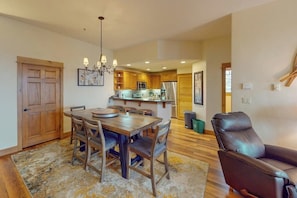 A dining area with a wooden table and chairs, a chandelier, and a kitchen in the background. - Walk-to-lift condo w/ shared hot tubs/pool/gym & amazing mountain views (Steamboat Springs)