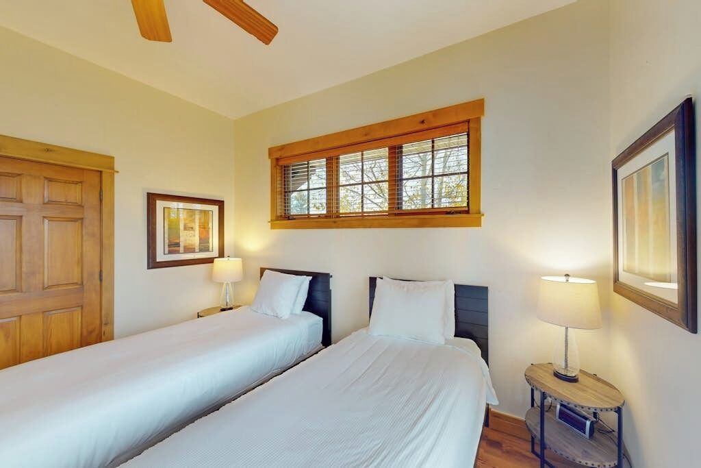A room with two beds, a wooden door, a window with blinds, a framed picture, and a small table with a lamp.