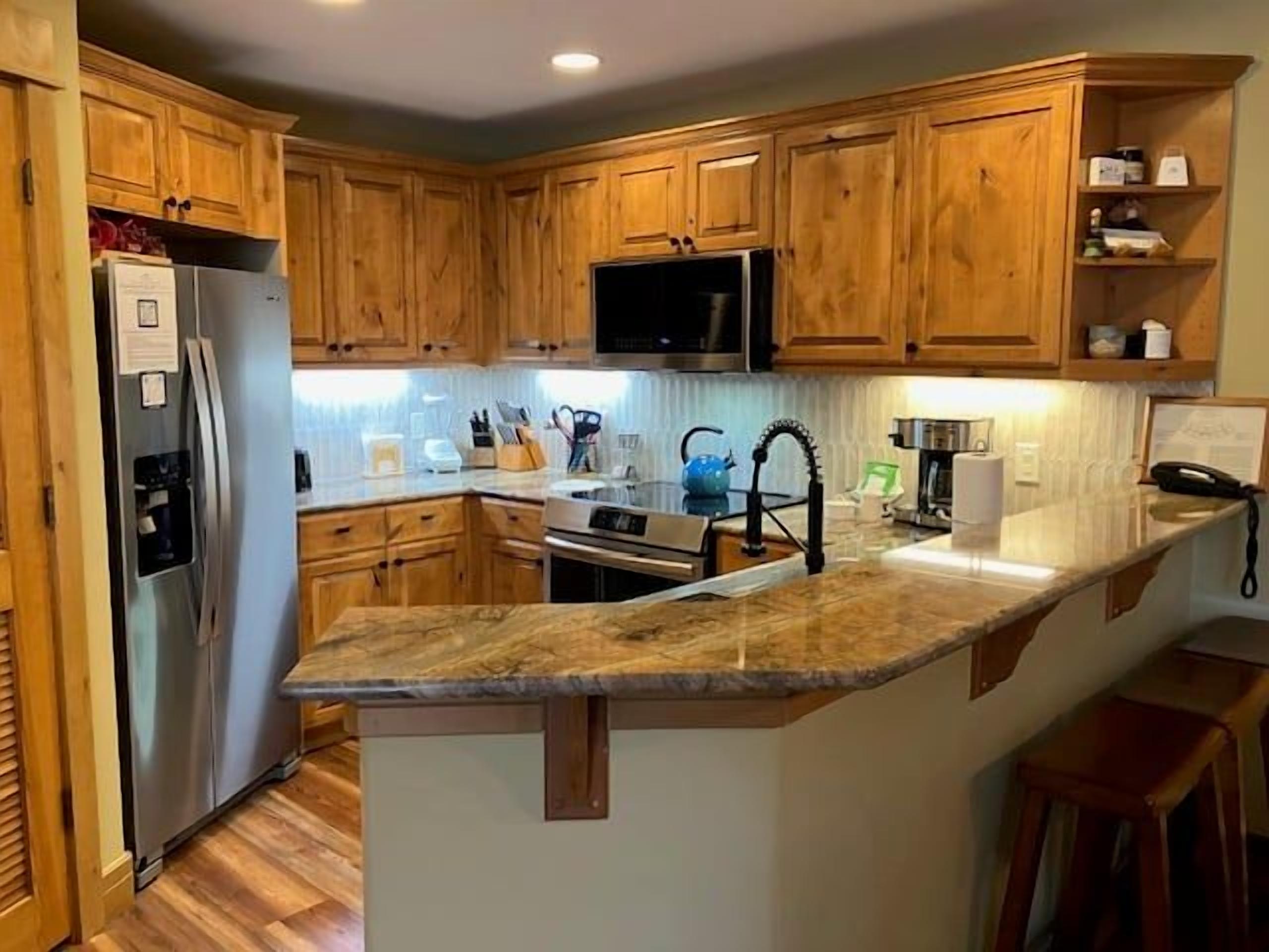 A kitchen with wooden cabinets, a stainless steel refrigerator, a microwave, a sink, and a granite countertop.