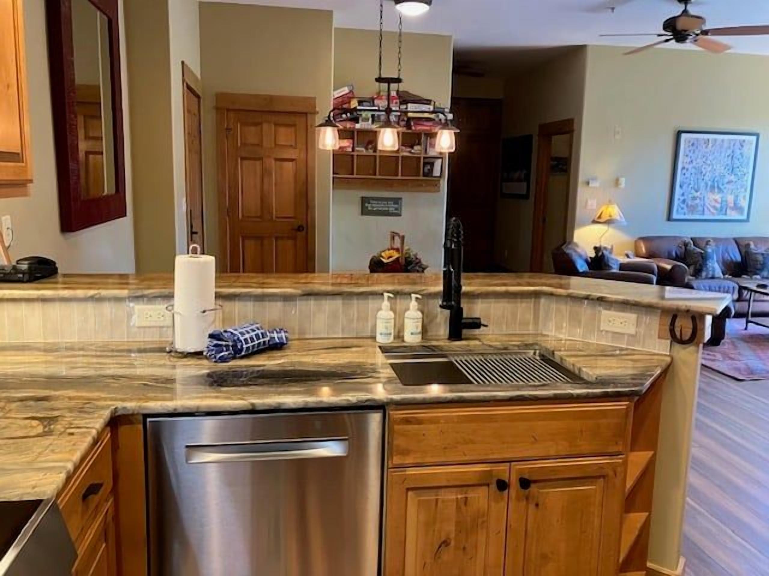 A kitchen with a stainless steel dishwasher, wooden cabinets, a marble countertop, and a hanging light fixture.