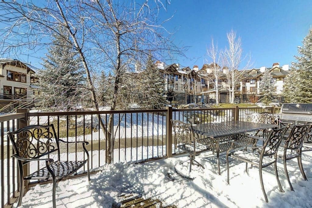 A snow-covered outdoor area with a metal table and chairs, a metal fence, and buildings in the background.