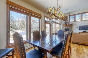 A dining area with a large wooden table, chairs with leather seats, a chandelier, and large windows with a view of trees. - EagleRidge Townhomes #1496 with central A/C, garage and private deck (Steamboat Springs)