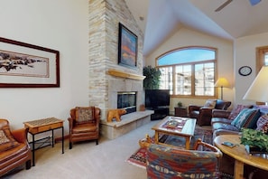 A living room with a stone fireplace, leather furniture, a large window, and a television. - Pretty & bright townhome in a great location for skiing w/ shared hot tubs (Steamboat Springs)