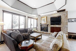 A living room with a brick fireplace, a large flat-screen TV, and a sofa set. - Ultimate mountain penthouse with hot tub, fitness, pool, & winter shuttle (Steamboat Springs)