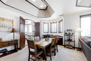 A dining area with a wooden table and chairs, a kitchen in the background, and a window with a view. - Ultimate mountain penthouse with hot tub, fitness, pool, & winter shuttle (Steamboat Springs)