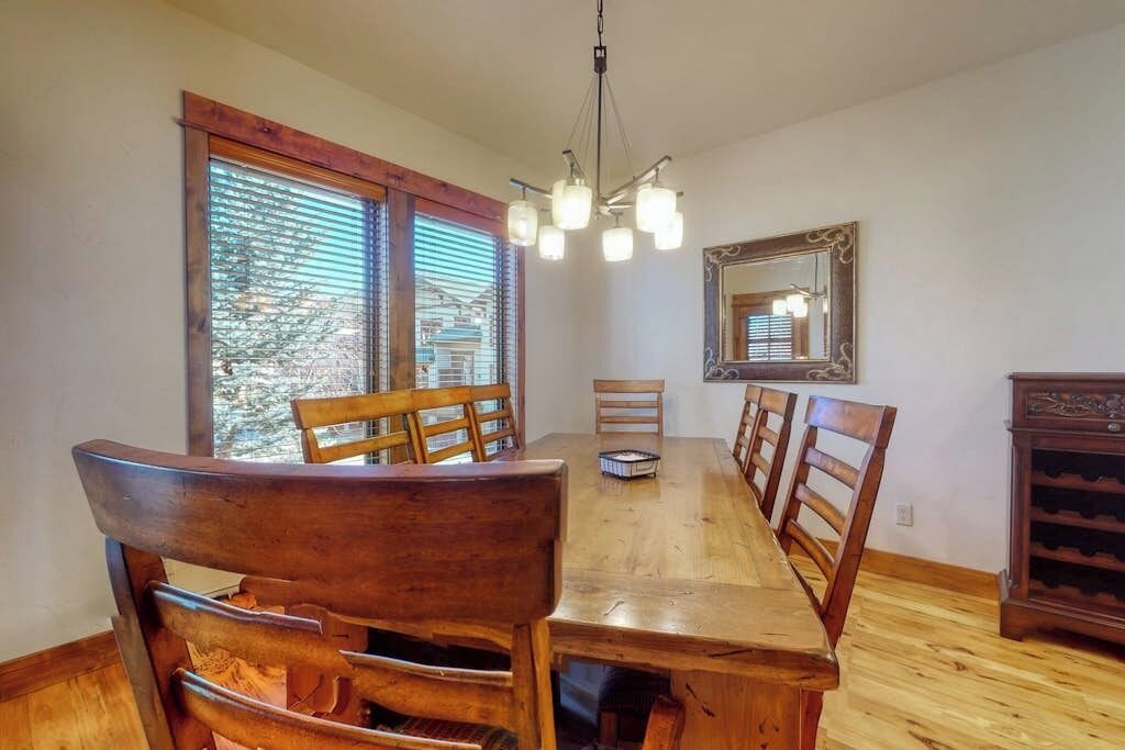 A dining area with a wooden table, chairs, a chandelier, and a large window with blinds.