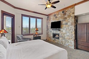 Ein Schlafzimmer mit Kamin aus Stein, Deckenventilator, Wand-Fernseher und Bett.