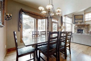 A dining area with a wooden table and chairs, a chandelier, and large windows. - 1516 EagleRidge Walk-to-gondola townhome w/ private hot tub & resort pool/firepit/hot tubs (Steamboat Springs)