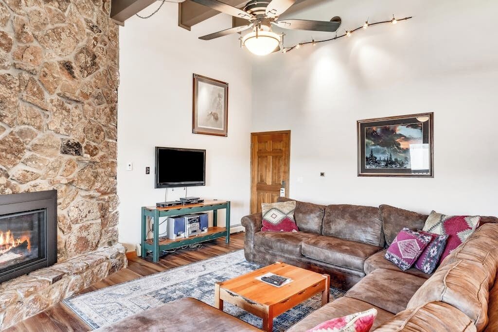 A living room with a stone wall, a fireplace, a leather sofa, a coffee table, a television, and a ceiling fan.