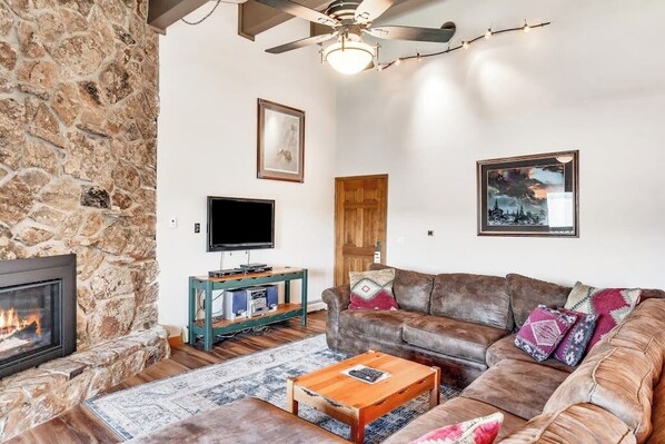 Una sala de estar con pared de piedra, chimenea, sofá de cuero, mesa de centro, televisor y ventilador de techo.