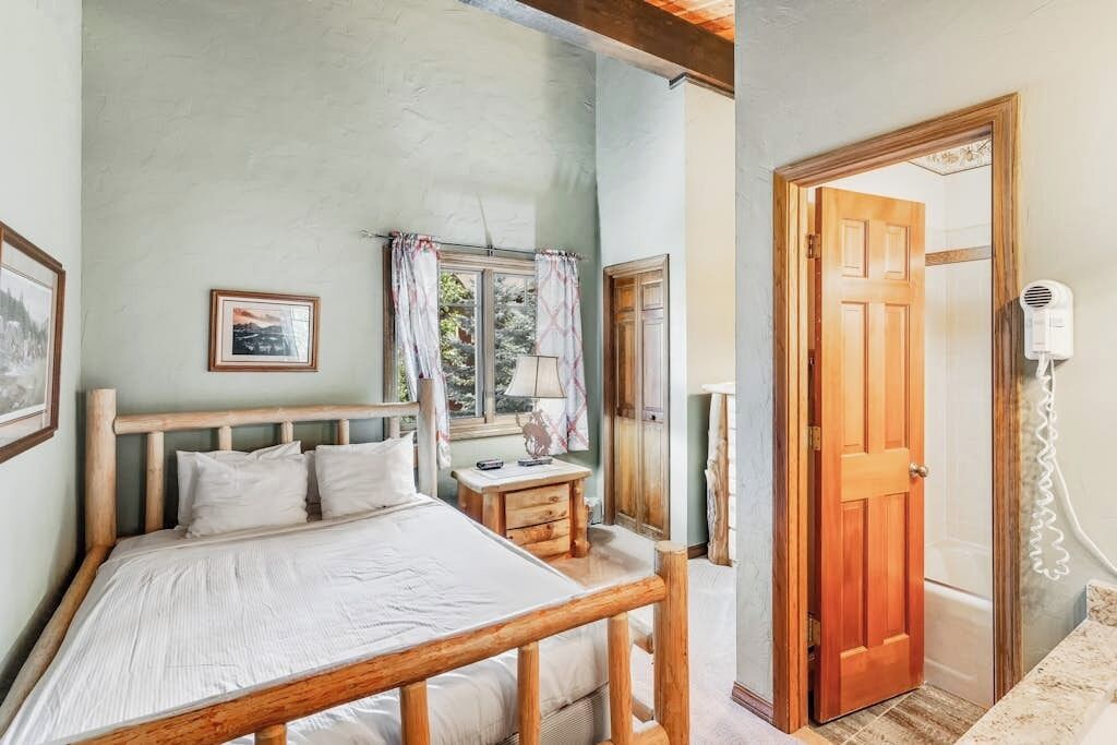 A bedroom with a wooden bed, a nightstand, a window with curtains, a door, and a hairdryer on the wall.