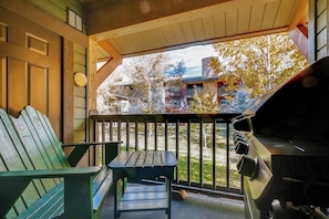 Eine Veranda mit Grill, zwei grüne Adirondack-Stühle und Blick auf ein Gebäude mit Bäumen.