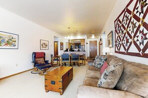 Ein Wohnzimmer mit einer Couch, Sesseln, einem Couchtisch und einer Holzkiste. Im Hintergrund ist eine Küche zu sehen.