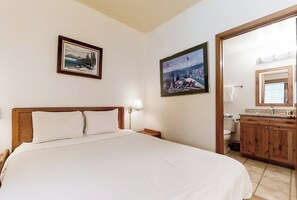 Ein Schlafzimmer mit Bett, Nachttisch, Wandlampe, Holz-Kopfteil und einem gerahmten Bild. Das Badezimmer verfügt über ein Waschbecken, einen Spiegel und eine Toilette.