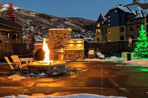 Ein Steinkamin mit Grill, umgeben von Schnee, in einem Innenhof mit Gebäuden und einem Weihnachtsbaum im Hintergrund.