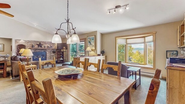 Uma sala de jantar ampla com mesa e cadeiras de madeira, lareira de tijolos e grandes janelas com vista para o exterior.