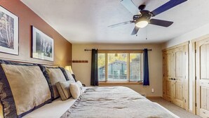 Quarto com cama, ventilador de teto, janela com cortinas e portas de madeira.