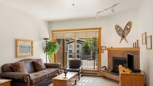 Ein Wohnzimmer mit Sofa, Sessel, Couchtisch, Kamin und einem großen Fenster mit Blick auf Gebäude und Bäume.