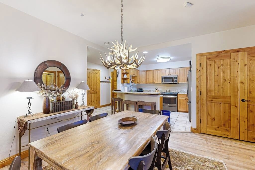 A dining area with a wooden table, chairs, a chandelier, and a kitchen in the background.