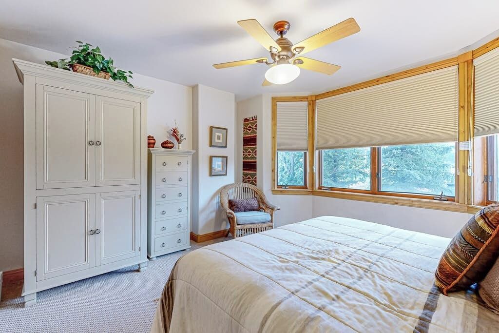 A bedroom with a bed, a dresser, a chair, a ceiling fan, and windows.