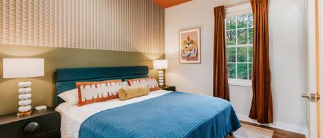 A bedroom with a bed, nightstands, a window with curtains, and a wall with blinds.