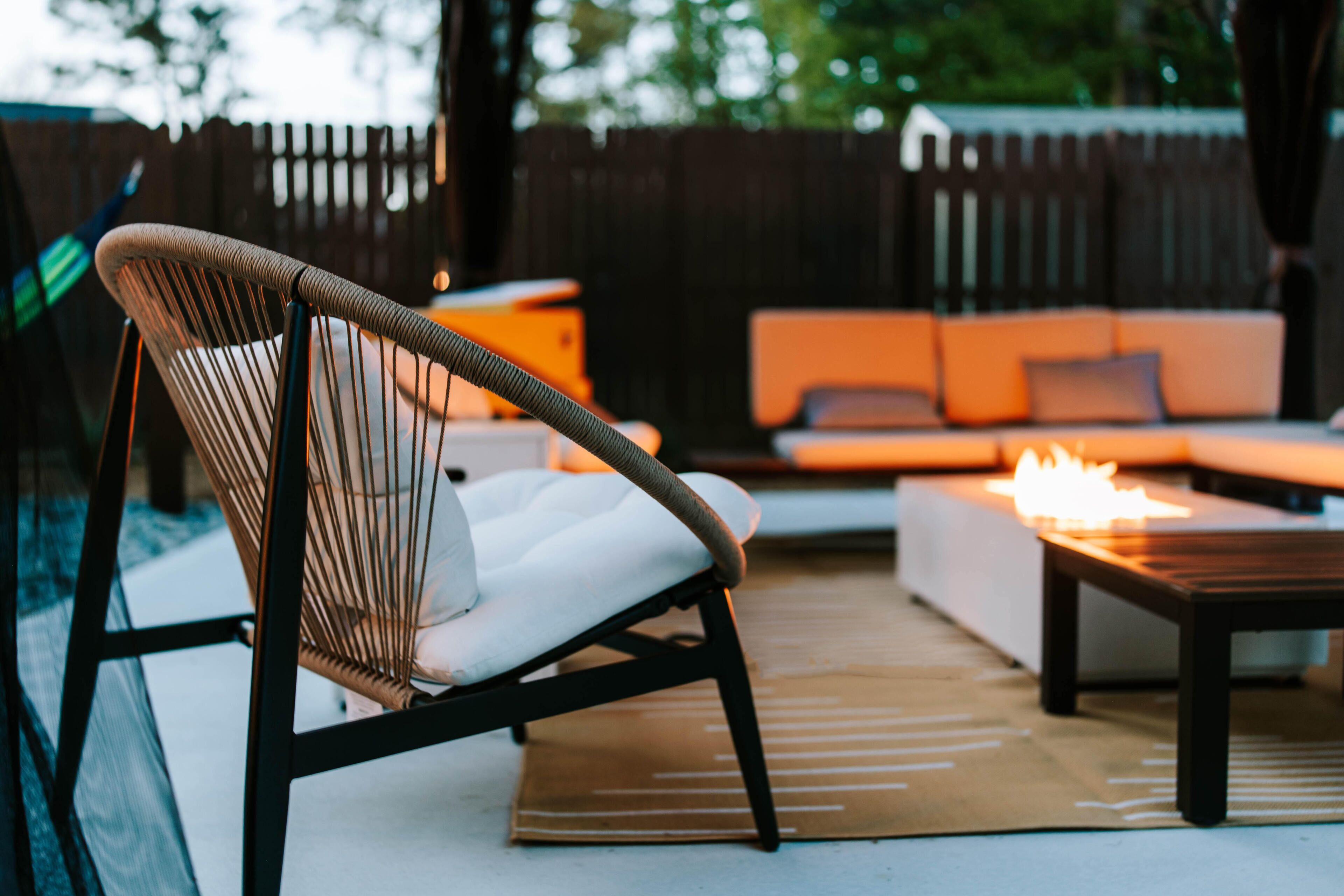 Une chaise en osier avec un coussin, un canapé, une petite table et un foyer sont disposés dans un espace extérieur.