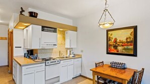 A compact kitchen with white cabinets, a wooden dining table, and a painting on the wall. - Base area studio w/shared pool & hot tub, private balcony - steps from ski lifts (Steamboat Springs)