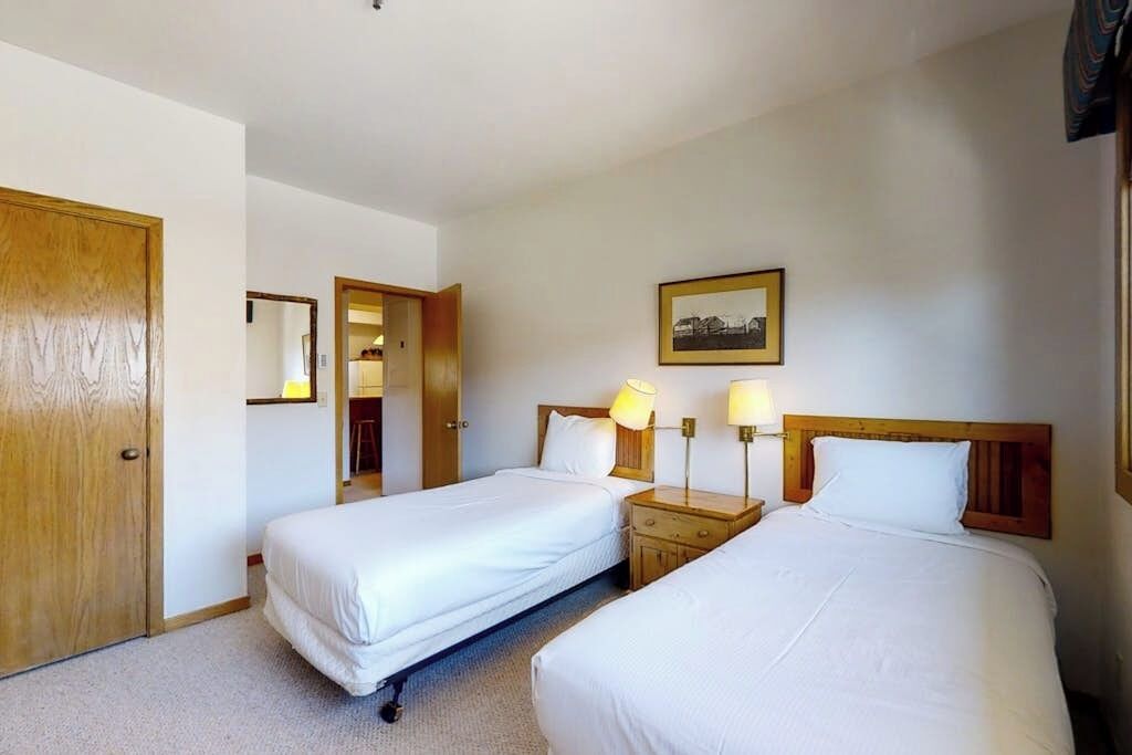 Habitación de hotel con dos camas individuales, un armario de madera, una mesita de noche con lámparas y un cuadro enmarcado en la pared.