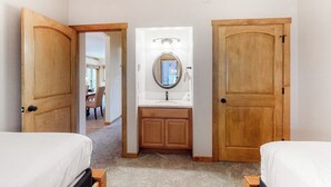Un dormitorio con dos camas, una puerta de madera, un baño con lavamanos y espejo, y un pasillo que conduce a otra habitación.