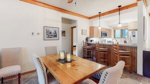 Un comedor con mesa y sillas de madera, un ventilador de techo y una cocina al fondo.