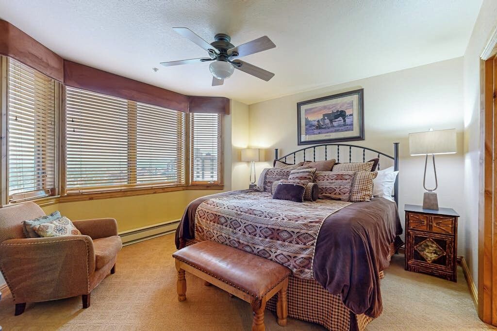 A bedroom with a bed, a chair, a bench, a ceiling fan, a window with blinds, a nightstand, and a lamp.