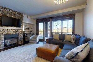 A living room with a stone fireplace, a flat-screen TV, a sofa, and a large window. - Stylish luxury condo w/gas fireplace, washer/dryer, shared pool, hot tubs, & gym (Steamboat Springs)