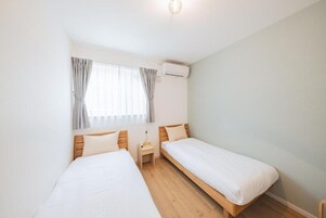 Una habitación pequeña y limpia con dos camas individuales, una mesita de noche, una ventana con cortinas y un equipo de aire acondicionado.