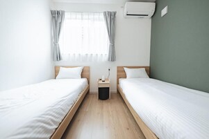 A room with two beds, a window with curtains, a small bedside table, and an air conditioning unit.