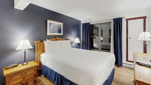 A hotel room with a bed, two bedside lamps, a wooden nightstand, a framed picture on the wall, a sliding glass door with curtains, and a wooden door. - Timber Run 416 - Appealing condo with sauna, pool, hot tubs & barbecue area (Steamboat Springs)