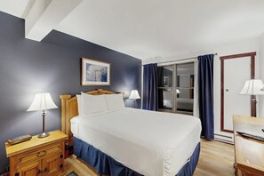 A hotel room with a bed, two bedside lamps, a wooden nightstand, a framed picture on the wall, a sliding glass door with curtains, and a wooden door. - Timber Run 416 - Appealing condo with sauna, pool, hot tubs & barbecue area (Steamboat Springs)