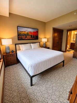 A hotel room with a large bed, two bedside tables with lamps, a painting on the wall, and a wooden door. - Trailhead Lodge #3107 condo with bonus den and gas fireplace (Steamboat Springs)