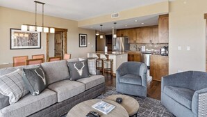 Een moderne woonkamer met een grijze bank, een ronde salontafel en een grijze fauteuil. Er is een keuken met houten kastjes en roestvrijstalen apparatuur.