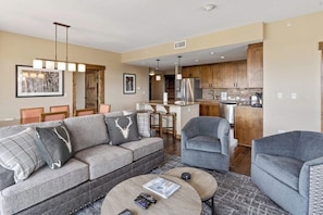 Ein modernes Wohnzimmer mit einem grauen Sofa, einem runden Couchtisch und einem grauen Sessel. Es gibt eine Küchenzeile mit Holzschränken und Edelstahlgeräten.