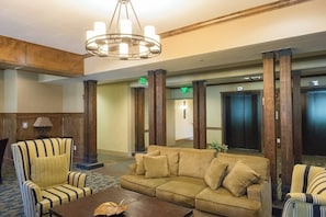 Eine Lobby mit einer Couch, Sesseln, einem Couchtisch, einer Deckenleuchte, Holzsäulen und einem Aufzug.