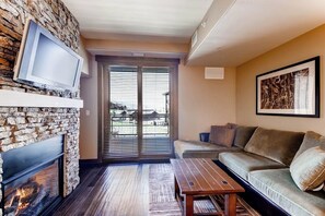 A living room with a stone fireplace, a flat-screen TV, a sofa, and a large window with blinds. - Beautiful studio w/private balcony & shared pool, hot tubs, fitness & game room (Steamboat Springs)